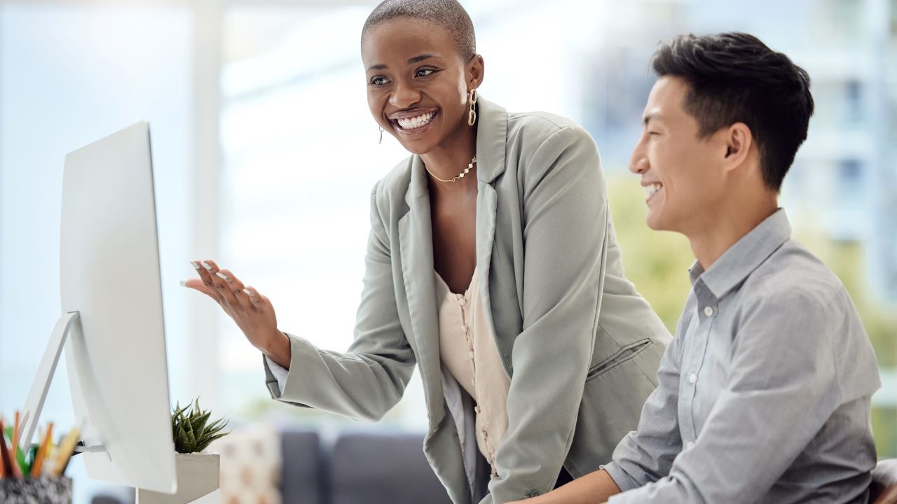 Young male employee seated at computer being trained by smiling female manager 